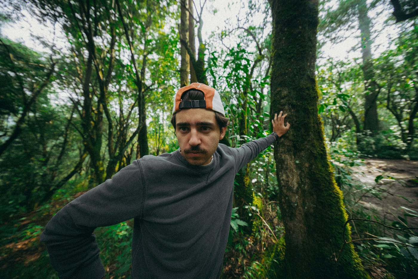 Recargado en un tronco con musgo, con la gorra puesta, mirando a la cámara
