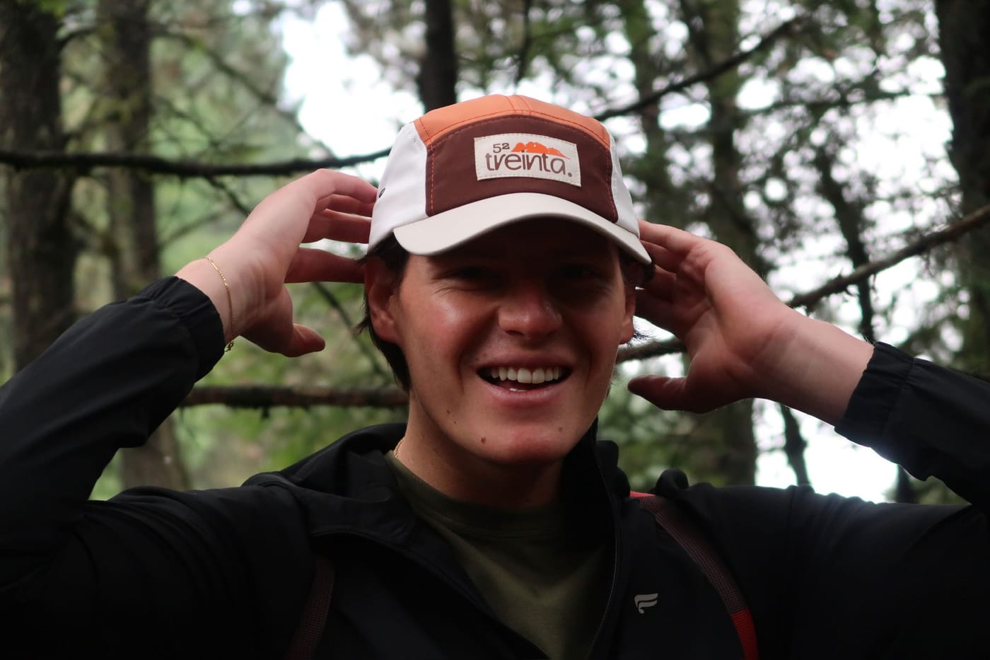 Sonriendo en el bosque mientras se acomoda la gorra con las dos manos