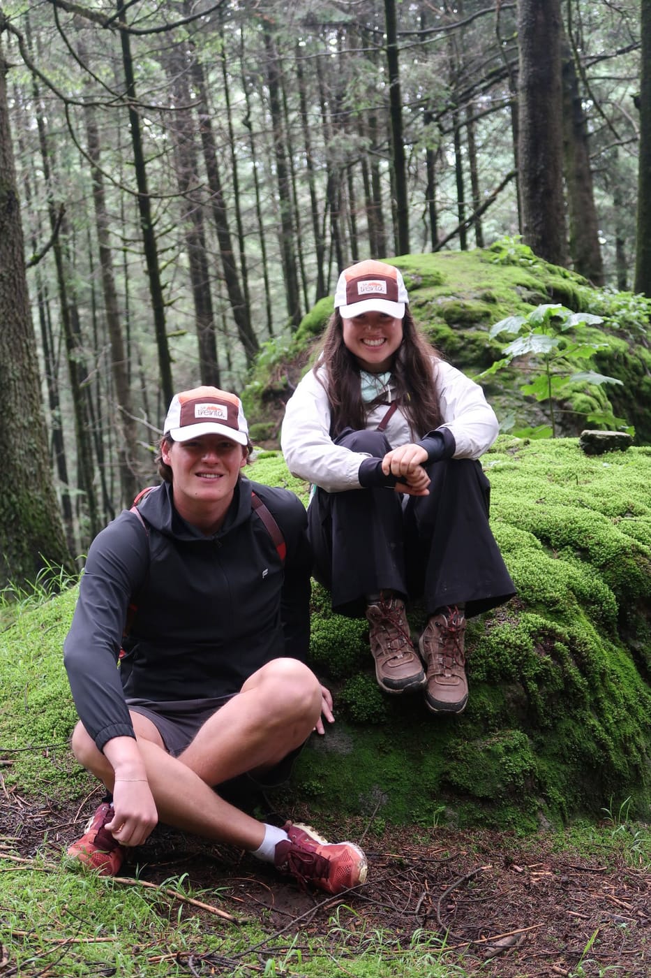 Dos personas sentadas en una roca con musgo en medio del bosque, las dos con la gorra