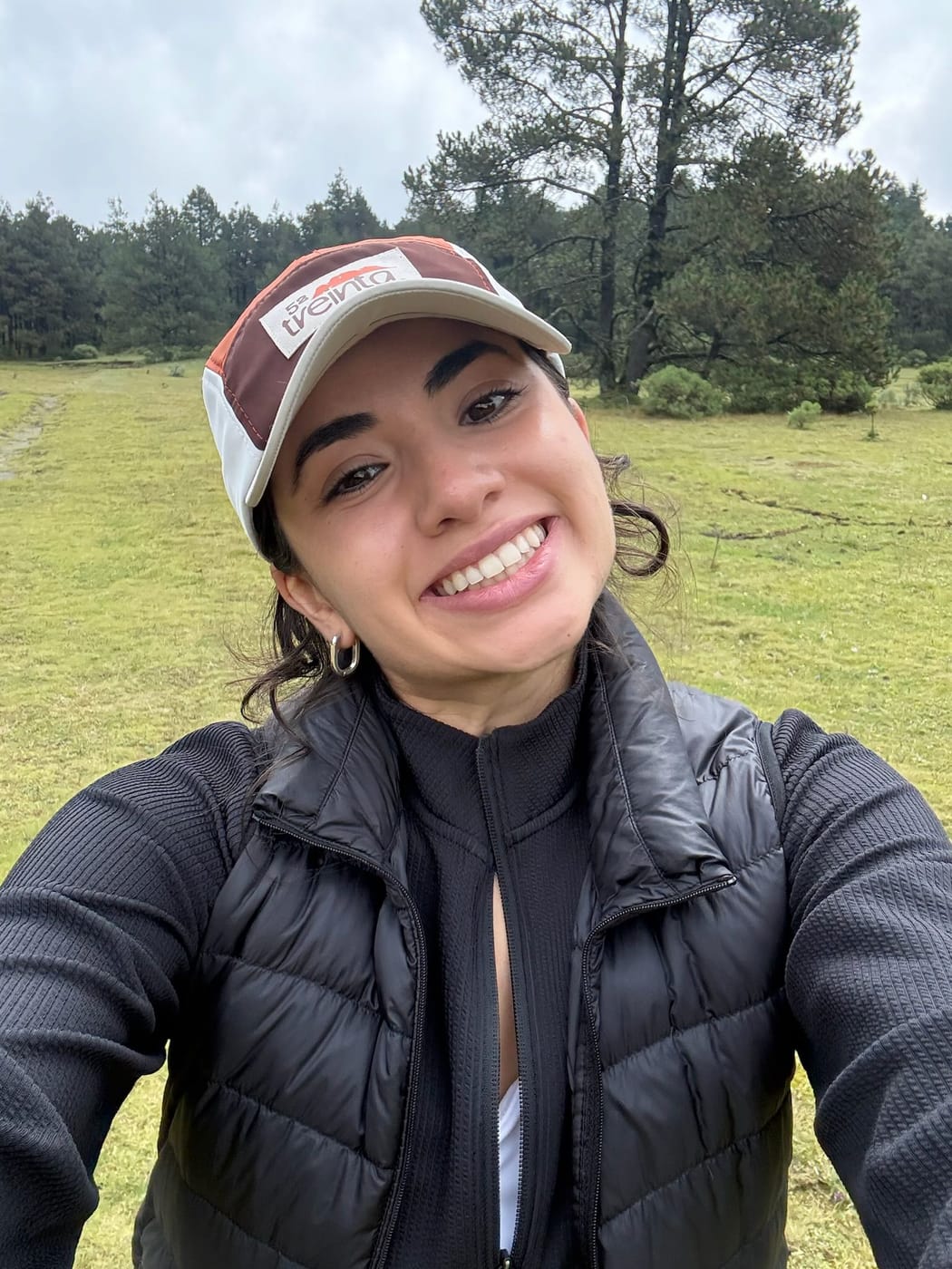 Una selfie en el pasto, sonriendo, con la gorra puesta y los pinos detrás