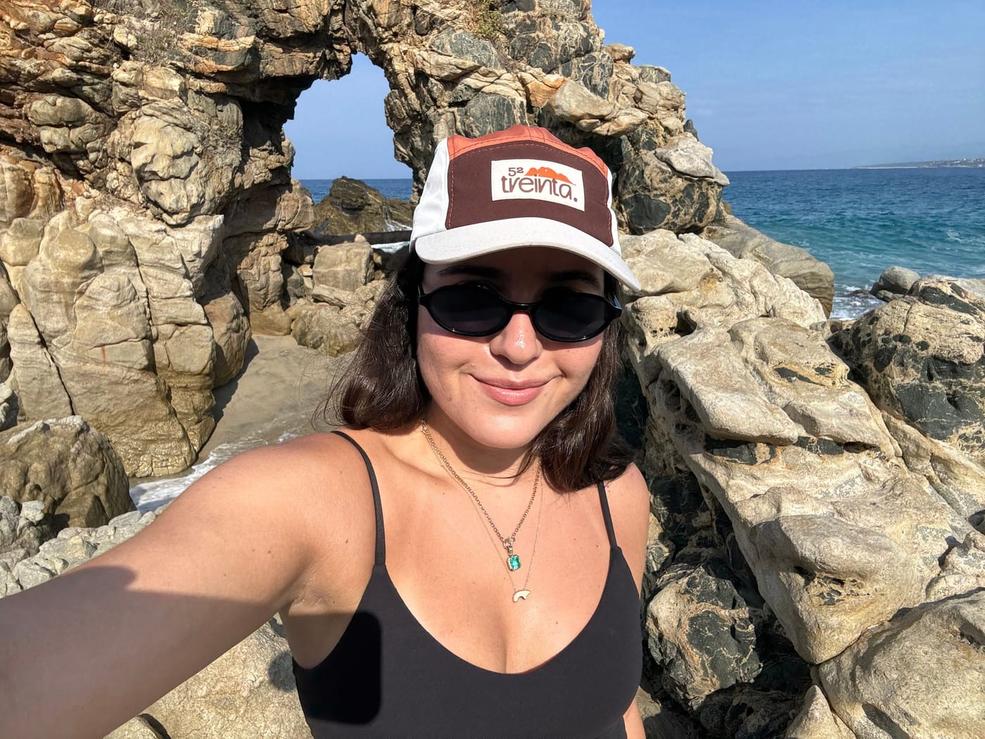 Una selfie en la costa, con lentes oscuros y la gorra puesta, frente a un arco de roca